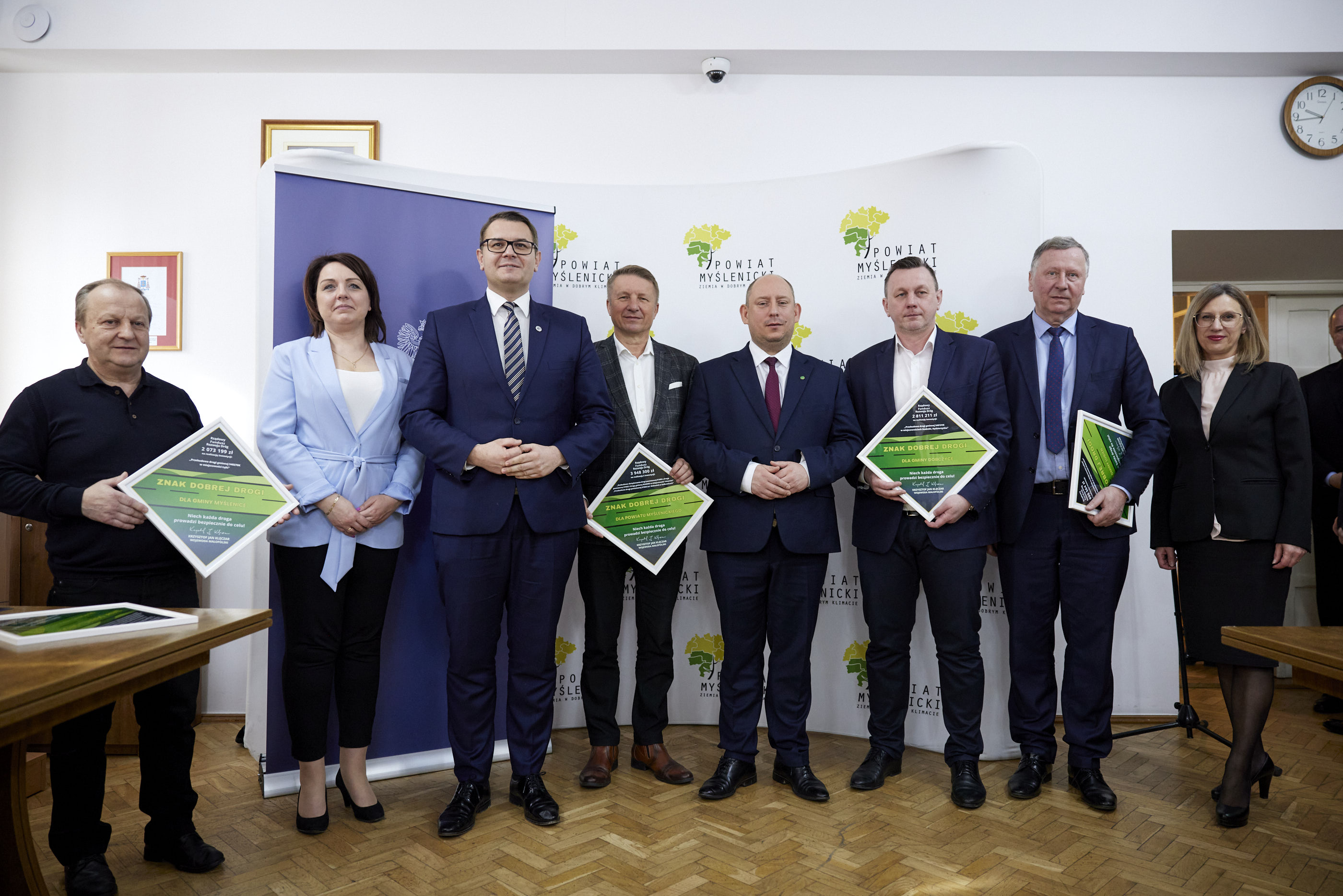 Wojewoda Krzysztof Jan Klęczar pozuje do zdjęcia z przedstawicielami samorządów z powiatu myślenickiego trzymającymi ramki z zieloną grafiką informującą o przyznanym dofinansowaniu.